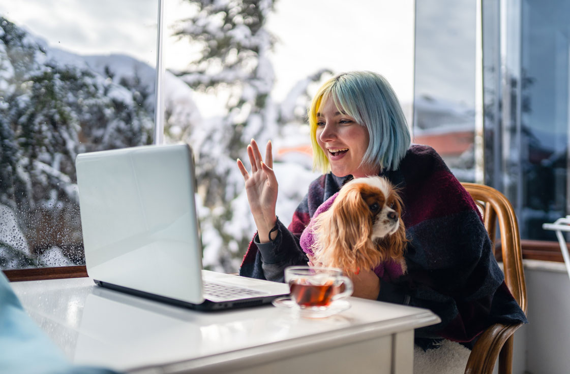 Junge Frau mit bunten Haaren sitzt mit Hund auf dem Schoss vor dem Laptop und winkt in die Kamera