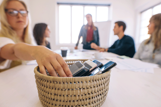 Mitarbeiter an einem Sitzungstisch legen ihr Smartphone in ein Körbchen, damit sie nicht abgelenkt werden