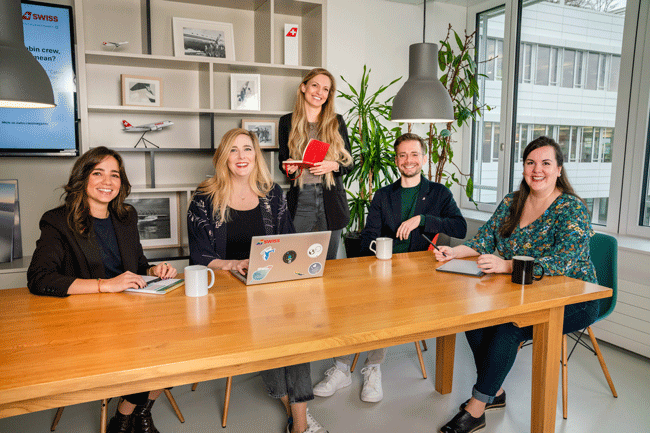 HR Team der Swiss International Airlines Maria Vega, Alana Gahler, Désirée Obrecht Gómez Rojas, Michael von Allmen und Isabel Silva (von links) sitzt zusammen an einem Holztisch in ihrem Büro