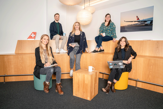 HR Team der Swiss International Airlines Maria Vega, Alana Gahler, Désirée Obrecht Gómez Rojas, Michael von Allmen und Isabel Silva (von links) sitzt zusammen an einem Holztisch in ihrem Büro