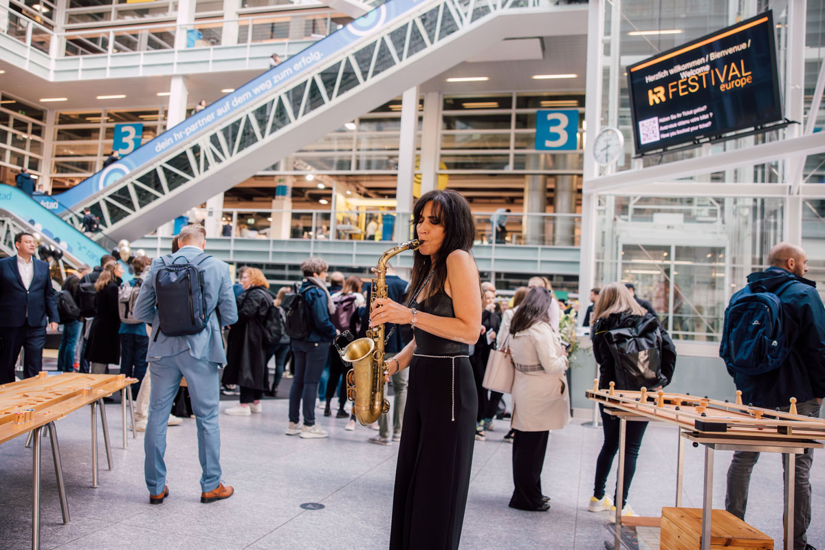 Saxophonistin spielt sanften Jazz in der Eingangshalle der Messe Zürich während Besuchende für den Eintritt Schlange stehen.