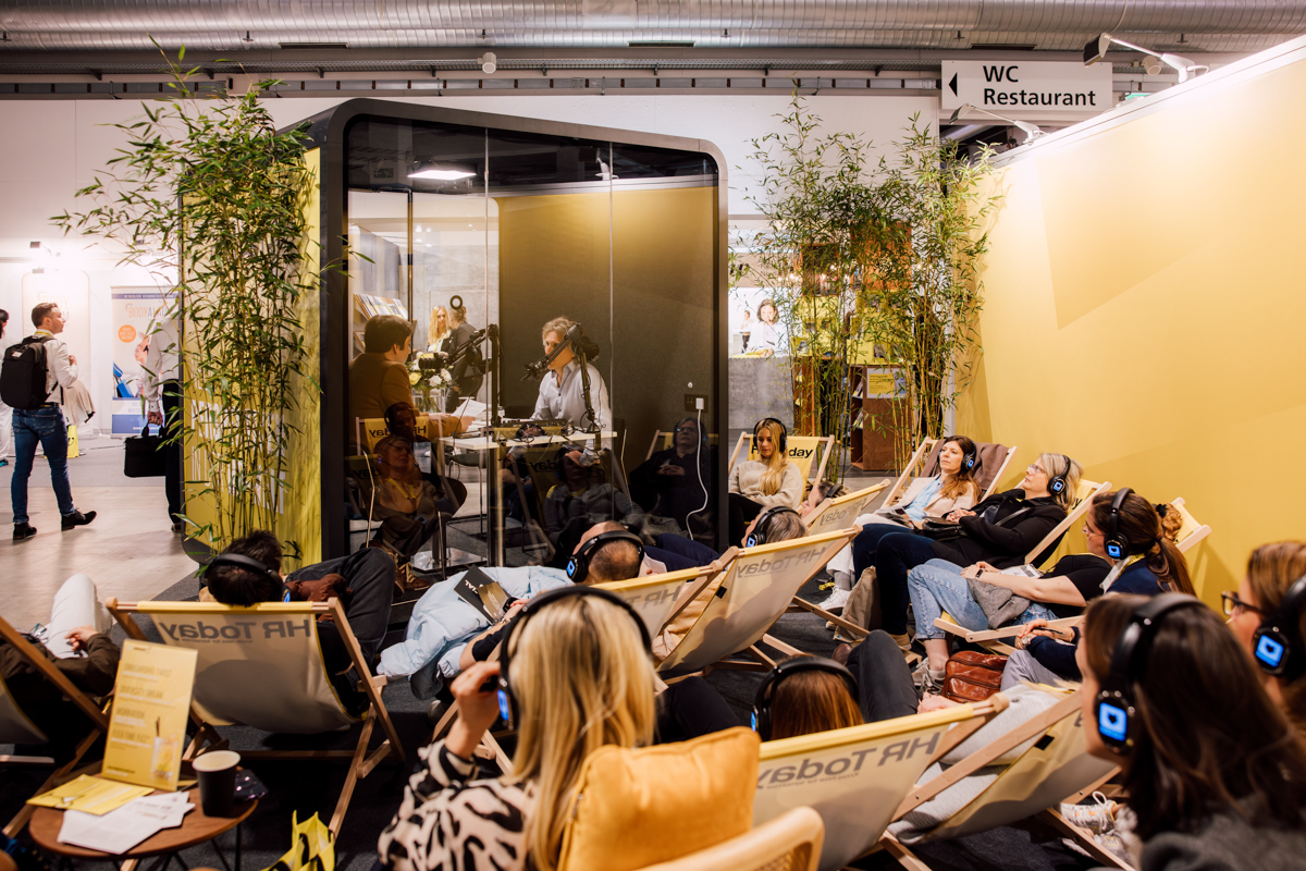 HR Today Stand am HR FESTIVAL europe. Chefredaktor Daniel Thüler sitzt im Booth mit Gast für die Live-Aufnahme des neuen Podcasts. Rundherum sitzen Zuhörerinnen und Zuhörer in Liegestühlen.