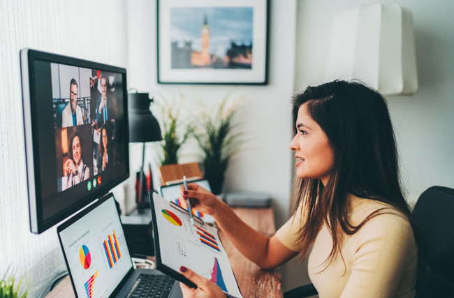 Frau arbeitet zu Hause im Home Office auf mehreren Bildschirmen, während sie an einer Video-Konferenz teilnimmt.