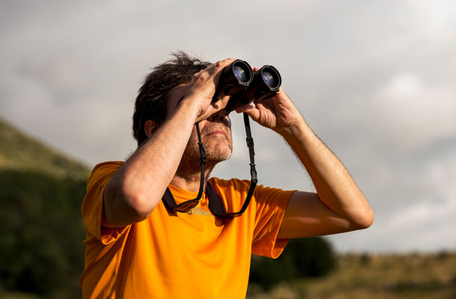 Ein Wanderer in orangen T-Shirt scannt mit Fernglas den Himmel nach Vögeln, symbolisch für wie Innovatoren in die Zukunft blicken.