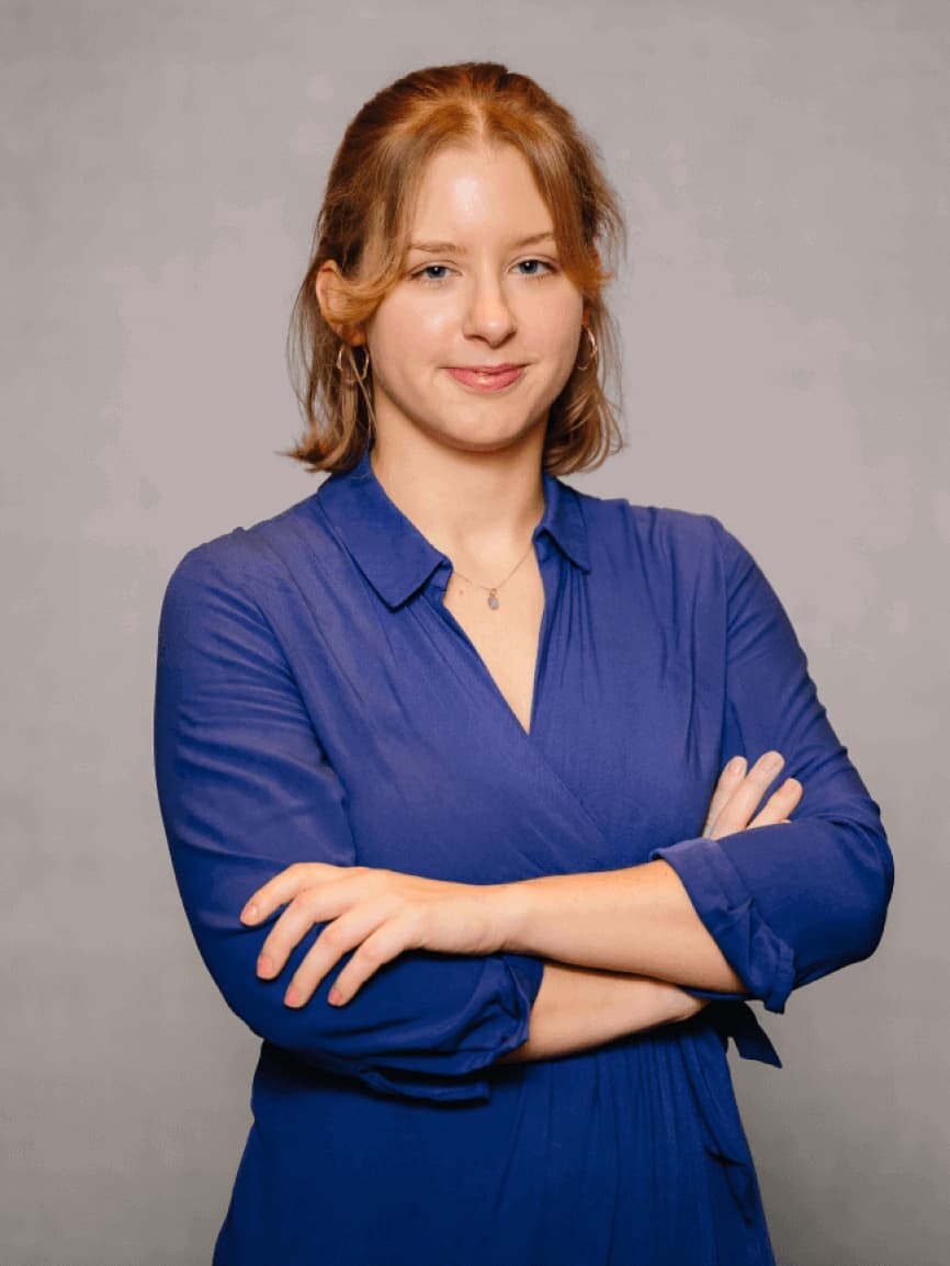 Das Bild zeigt die Autorin, eine junge Frau mit blauer Bluse und rostroten Haaren. Sie hat die Arme vor dem Körper verschränkt.