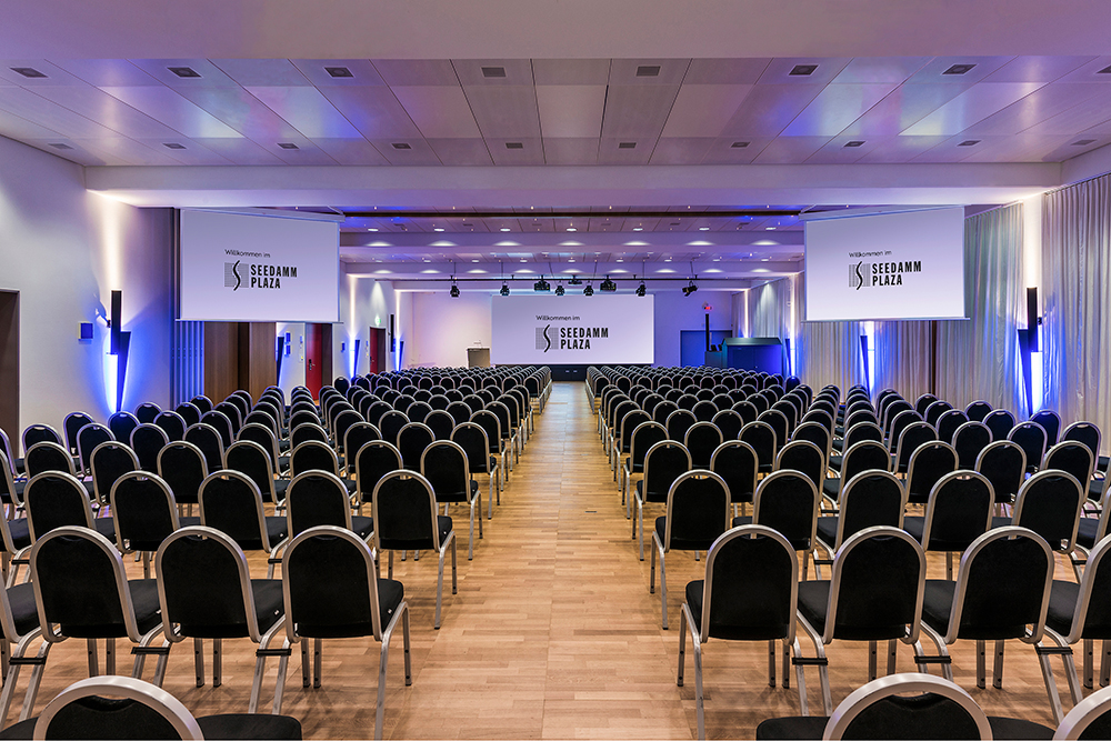 Innenaufnahme eines grossen Konferenzsaals im Seedamm Plaza, mit vielen gleichmässig angeordneten schwarzen Stühlen, einem Rednerpult auf der Bühne und drei grossen Projektionsflächen mit dem Logo des Hotels, in blauer Ambientebeleuchtung.