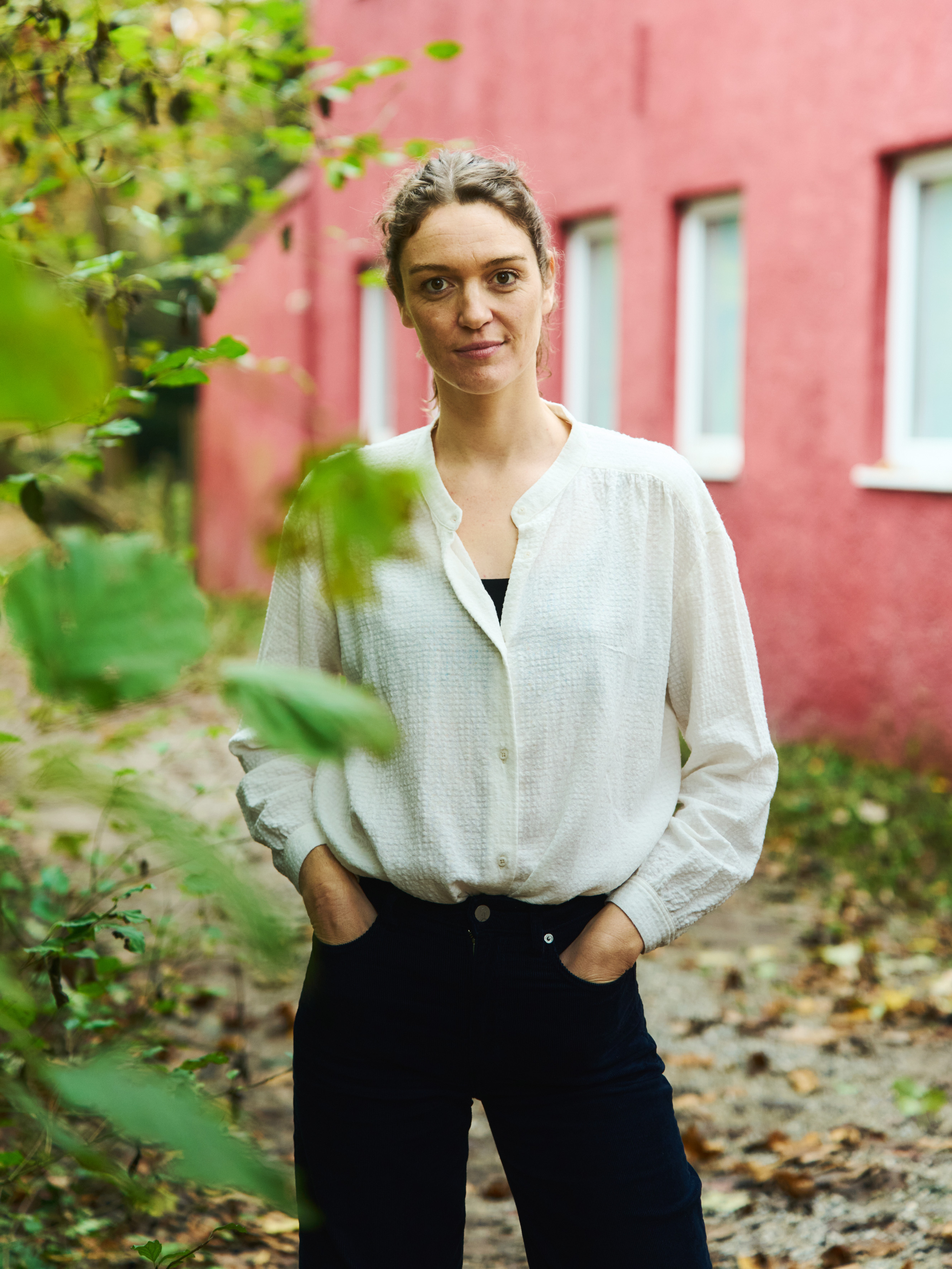 Porträt von Elisabeth Raes, einer Frau, die im Freien auf einem Laubweg vor einem roten Gebäude mit weissen Fenstern steht; sie trägt eine weisse Bluse und eine dunkle Hose und blickt ruhig in die Kamera, umgeben von grünem Blattwerk.
