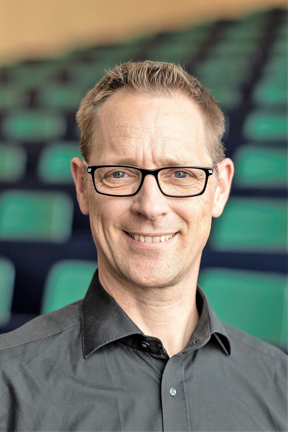 Porträt eines Mannes mit kurzem, hellbraunem Haar und Brille, der freundlich in die Kamera lächelt. Er trägt ein dunkles Hemd und steht vor einer Sitzreihe mit grün-schwarzen Stühlen, vermutlich in einem Hörsaal.  