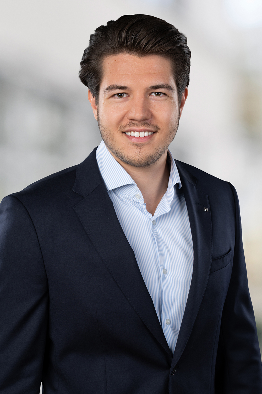 Porträt eines jungen Mannes mit dunklem, zurückgekämmtem Haar, der lächelnd in die Kamera schaut. Er trägt ein dunkelblaues Sakko über einem hellblauen Hemd. Der Hintergrund ist ein heller, unscharfer Büro- oder Gebäudeflur.