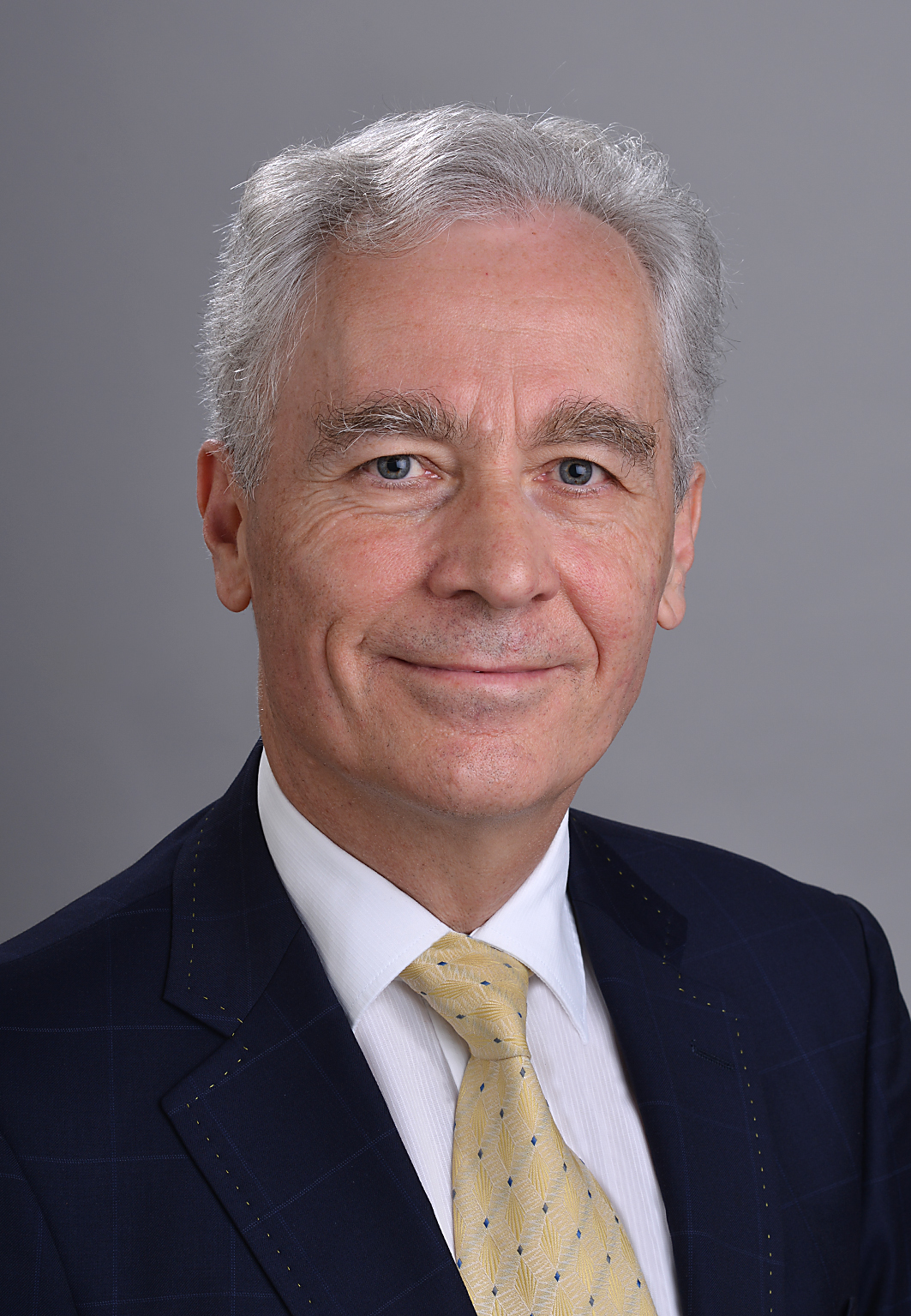 Porträt eines älteren Mannes mit grauen Haaren und hellem Teint, der direkt in die Kamera lächelt. Er trägt einen dunkelblauen Anzug mit feinem Karomuster, ein weisses Hemd und eine gelbe Krawatte mit dezentem Muster. Der Hintergrund ist neutral grau.