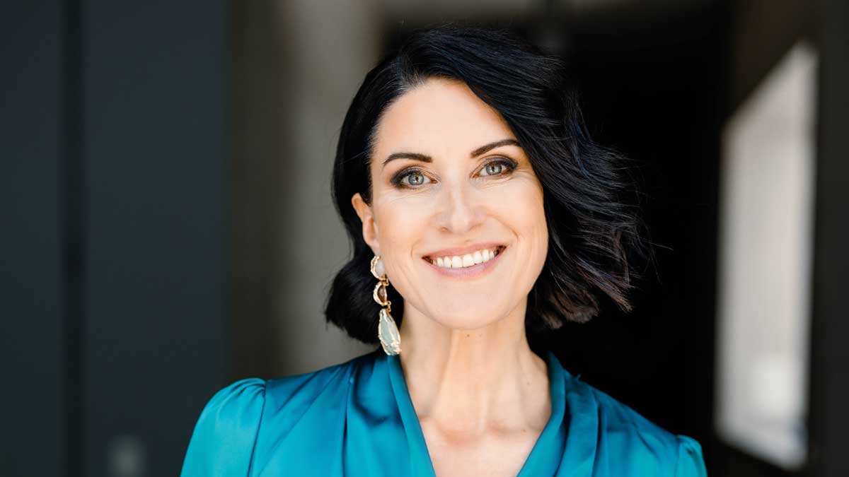 Porträt von Verena Kemperling, einer lächelnden Frau mit schulterlangen dunklen Haaren, die grosse Ohrringe und eine türkisfarbene Bluse trägt; der Hintergrund ist unscharf und dunkel gehalten.