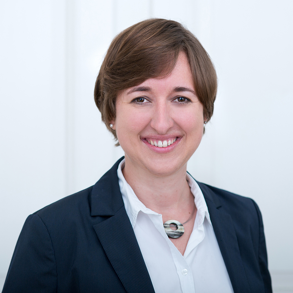 Porträt einer lächelnden Frau mit kurzen braunhaarigen Haaren. Sie trägt ein dunkelblaues Blazer-Jackett über einer weissen Bluse und eine auffällige runde Halskette mit Marmormuster. Der Hintergrund ist schlicht und weiss.