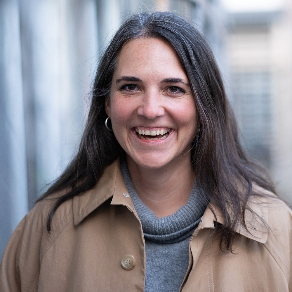 Porträt einer lachenden Frau mit langen dunkelbraunen Haaren, die eine beigefarbene Jacke und einen grauen Rollkragenpullover trägt; im Hintergrund eine unscharfe städtische Gasse mit Gebäuden.