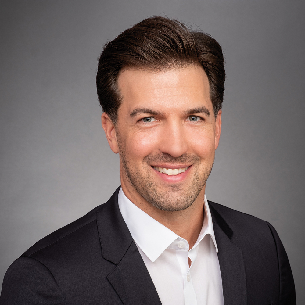 Porträt eines lächelnden Mannes mit dunklem, nach hinten frisiertem Haar und kurzem Bart. Er trägt ein dunkles Sakko über einem weissen Hemd und steht vor einem neutral grauen Hintergrund.