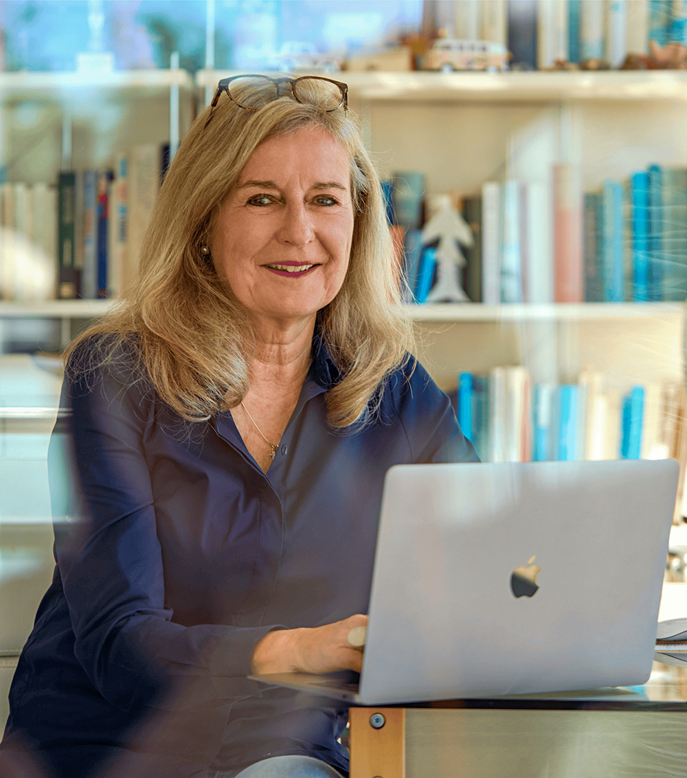 Ältere Frau mit langen blonden Haaren sitzt lächelnd an einem Tisch und arbeitet an einem silbernen MacBook-Laptop. Sie trägt eine dunkelblaue Bluse und hat eine Brille auf dem Kopf. Im Hintergrund ist ein Bücherregal mit vielen Büchern und Dekorationsobjekten zu sehen.