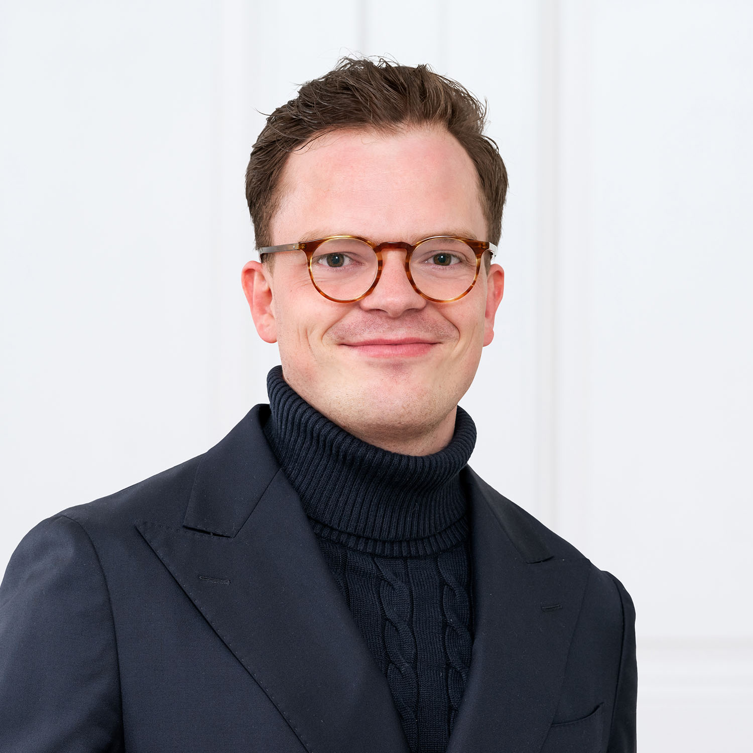 Porträt eines lächelnden Mannes mit kurzen braunen Haaren und runder Brille. Er trägt einen dunkelblauen Rollkragenpullover mit Zopfmuster unter einem dunkelblauen Anzugjackett. Der Hintergrund ist schlicht und weiss.