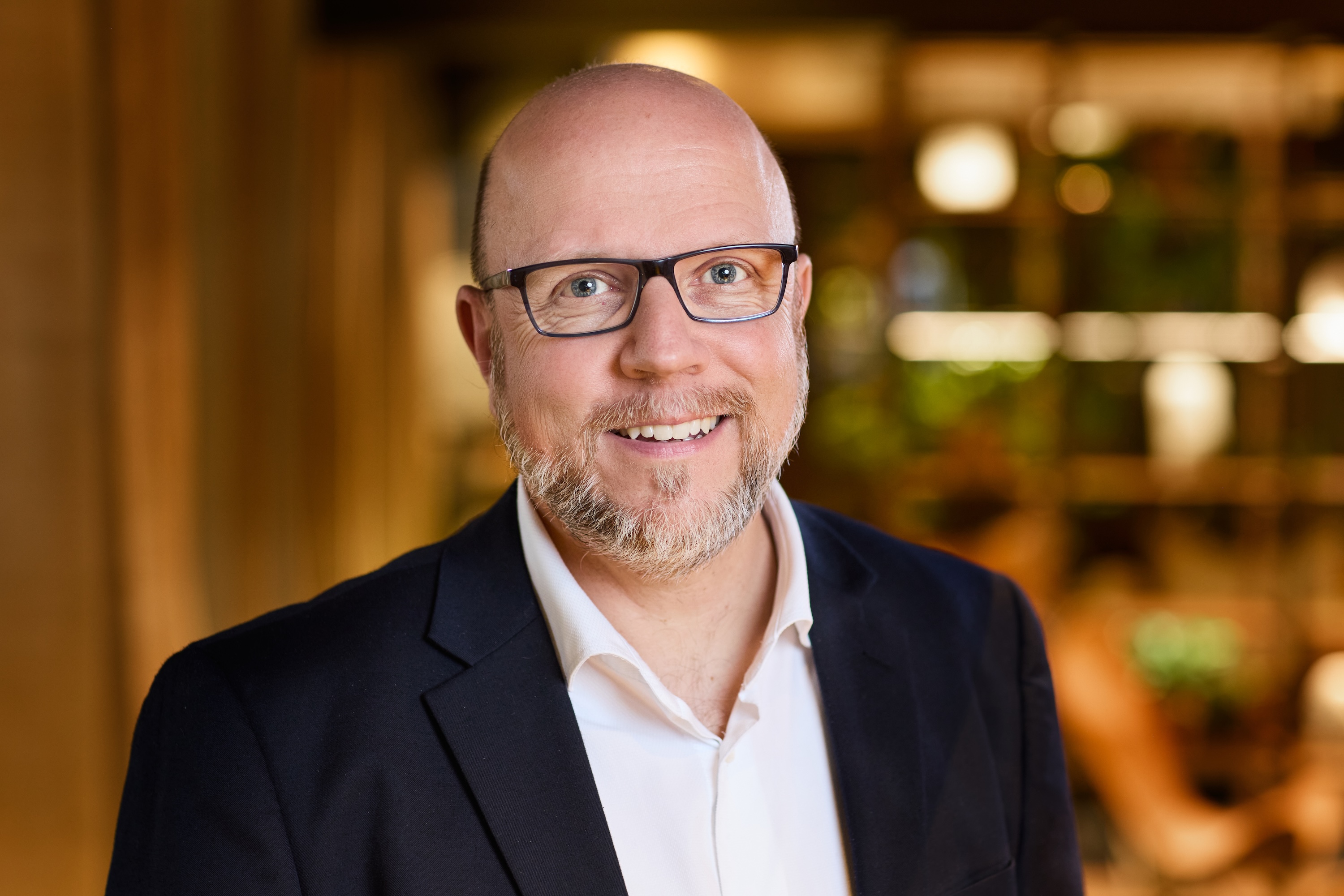 Porträt von Ralf Räber, der freundlich in die Kamera lächelt. Er trägt eine Brille mit dunklem Gestell, einen gepflegten graumelierten Bart und ist mit einem dunklen Sakko über einem weißen Hemd bekleidet. Der Hintergrund ist in warmen, weichen Lichttönen unscharf gehalten.