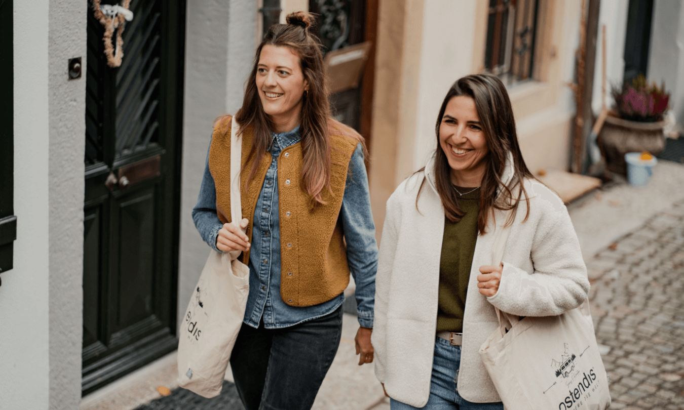 Zwei lachende Frauen spazieren nebeneinander. Die Frau auf der linken Seite hat lange braune Haare, die teilweise zu einem Dutt hochgesteckt sind. Sie trägt ein blaues Jeanshemd unter einer braun-senffarbenen Teddyfell-Weste und eine dunkle Hose. Die Frau rechts hat ebenfalls lange, dunkle Haare und trägt einen olivgrünen Pullover, eine cremefarbene Teddyfell-Jacke sowie eine blaue Jeans. Beide Frauen tragen helle Stoffbeutel mit der Aufschrift und dem Logo "ostendis".