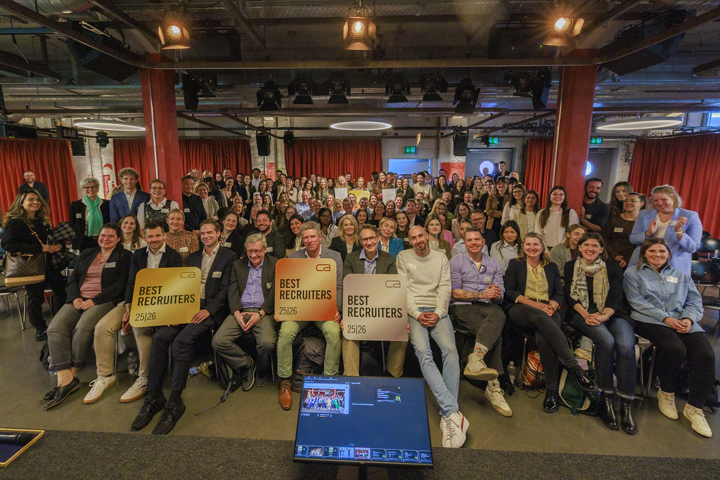 Eine große Gruppe von Menschen posiert für ein Gruppenfoto in einem Veranstaltungsraum im Industrie-Stil mit roten Vorhängen im Hintergrund. In der vorderen Reihe halten drei Personen große, quadratische Schilder mit der Aufschrift 'BEST RECRUITERS 25|26'. Im Vordergrund ist ein Monitor zu sehen, der die Aufnahmeumgebung eines Fotografen anzeigt.