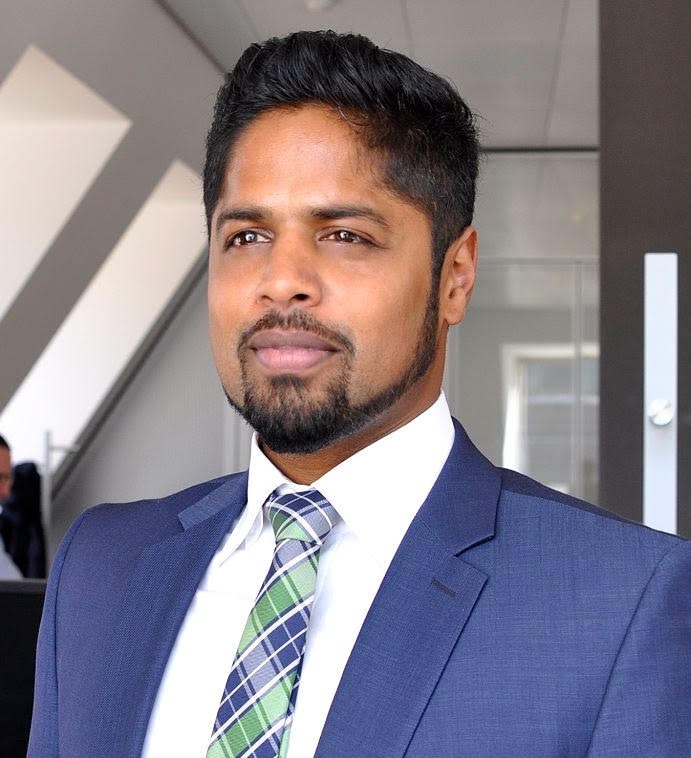 Ein Porträt eines Mannes mit dunklem Haar und Bart. Er trägt einen dunkelblauen Anzug, ein weißes Hemd und eine grün-blau karierte Krawatte. Er blickt mit neutralem, professionellem Ausdruck leicht an der Kamera vorbei nach links. Im unscharfen Hintergrund ist eine helle Büroumgebung zu erkennen.