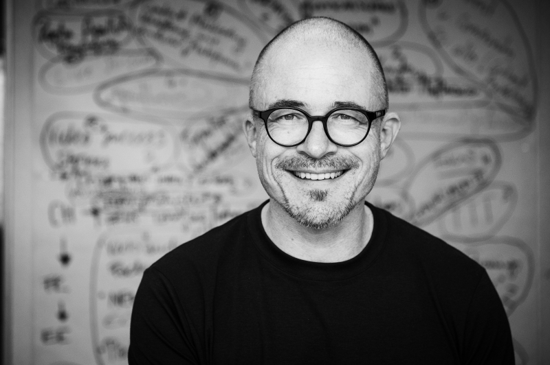 Schwarz-Weiß-Aufnahme eines Mannes, der freundlich in die Kamera blickt. Er hat eine Glatze, einen kurzen Kinnbart und trägt eine markante, runde Brille sowie ein schwarzes T-Shirt. Im unscharfen Hintergrund ist ein Whiteboard oder eine helle Wand zu erkennen, die mit zahlreichen handschriftlichen Notizen, umkreisten Begriffen und Skizzen gefüllt ist.
