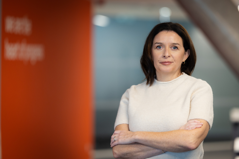 Porträt einer Frau mit dunklem, schulterlangem Haar in einem hellen, kurzärmligen Oberteil, die mit verschränkten Armen direkt in die Kamera blickt. Im unscharfen Hintergrund befindet sich links eine leuchtend orangefarbene Wand mit verschwommenem Text und rechts eine angedeutete Büroumgebung.