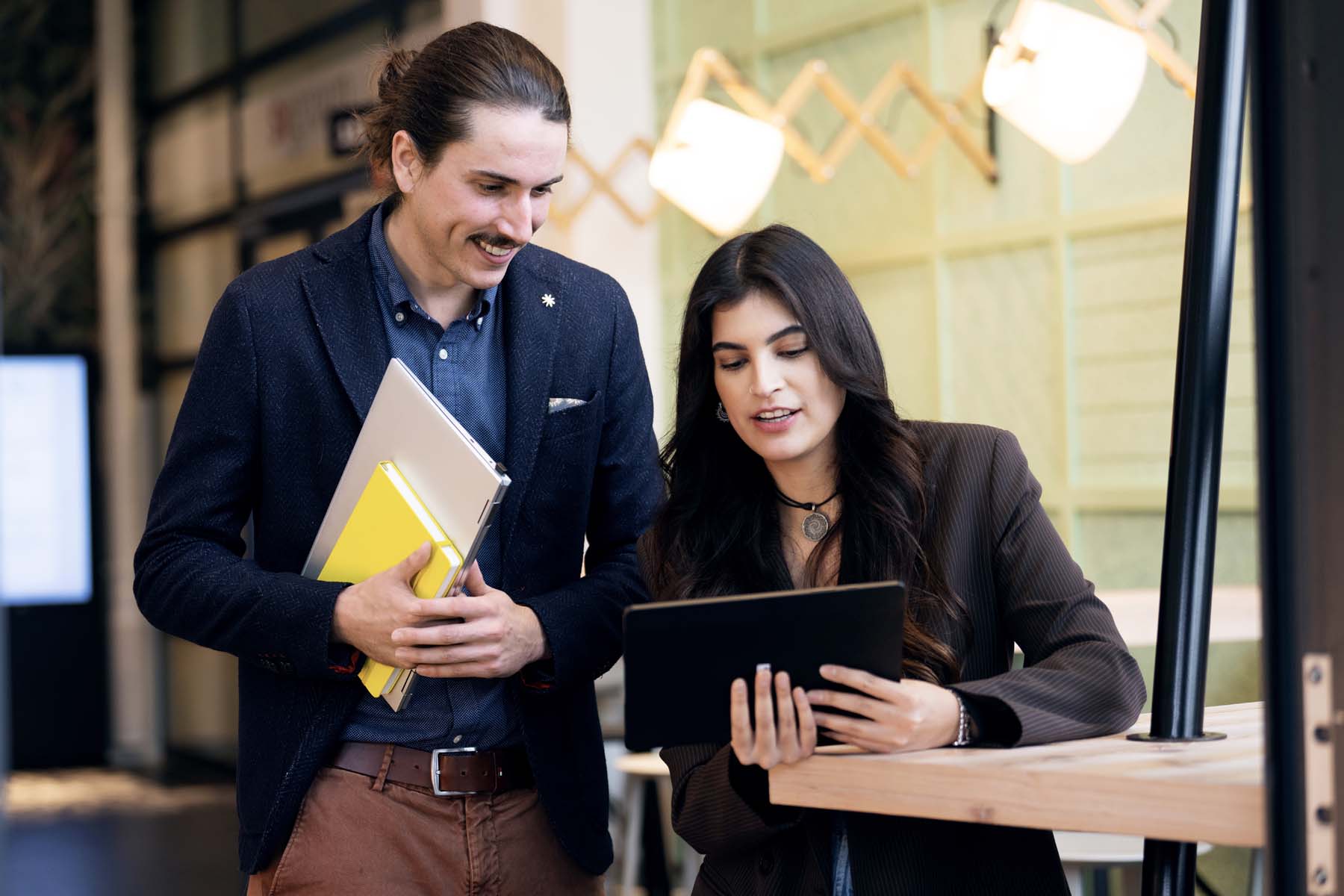 Eine Frau und ein Mann arbeiten in einer modern gestalteten Büroumgebung zusammen. Die Frau mit langen, dunklen Haaren trägt einen dunklen Nadelstreifen-Blazer, lehnt an einem hölzernen Stehtisch und hält ein Tablet, auf das beide blicken. Der Mann mit zusammengebundenem Haar und Schnurrbart steht lächelnd daneben; er trägt ein blaues Hemd unter einem dunklen Sakko und hält ein Laptop sowie ein auffälliges gelbes Notizbuch im Arm.
