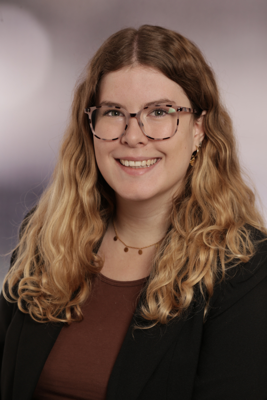 Gastautorin Alexa Ramgraber blickt freundlich lächelnd direkt in die Kamera. Sie trägt eine Brille mit gemustertem Rahmen, ein braunes Top unter einem schwarzen Blazer sowie eine feine Goldkette. Ihr langes, gewelltes Haar fällt offen über die Schultern vor einem neutralen Studiohintergrund.
