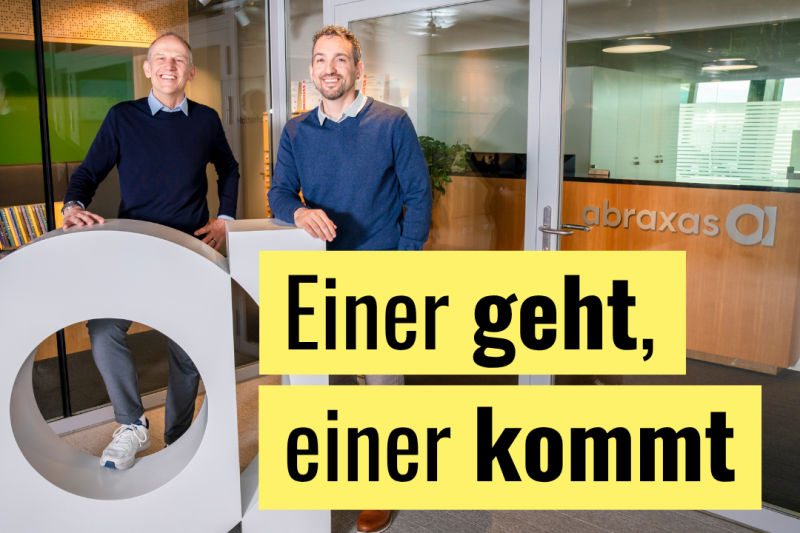 Redaktionelle Grafik mit dem Titeltext «Einer geht, einer kommt» auf gelbem Balken im unteren Bildbereich: Zwei lächelnde Männer mittleren Alters stehen im Eingangsbereich des Unternehmens Abraxas — erkennbar am Logo an der Glaswand im Hintergrund — und lehnen an einem weissen Objekt in Form des Abraxas-«a»-Logos.