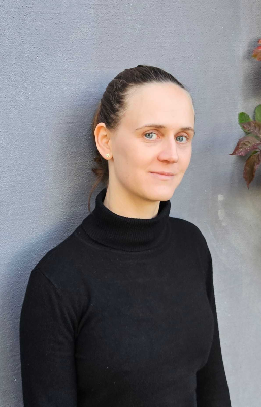 Porträt einer Frau mit dezentem Lächeln und zurückgebundenem Haar, die einen schwarzen Rollkragenpullover trägt. Sie steht vor einer grauen Wand, an deren rechtem Rand einige dunkle Blätter ins Bild ragen.