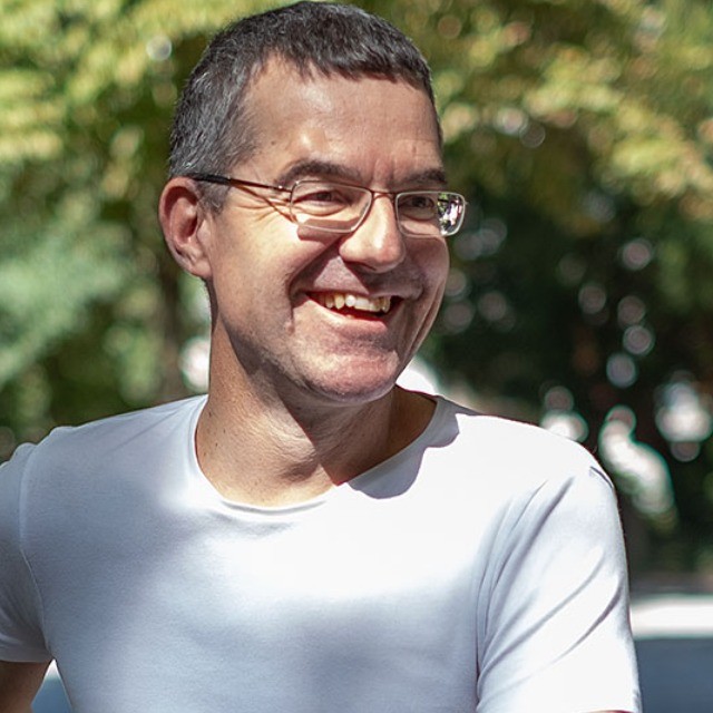 Ein freundlich lächelnder Mann mit Brille und kurzen, leicht ergrauten Haaren blickt leicht zur Seite. Er trägt ein weißes T-Shirt vor einem unscharfen, von Sonnenlicht durchfluteten grünen Hintergrund.