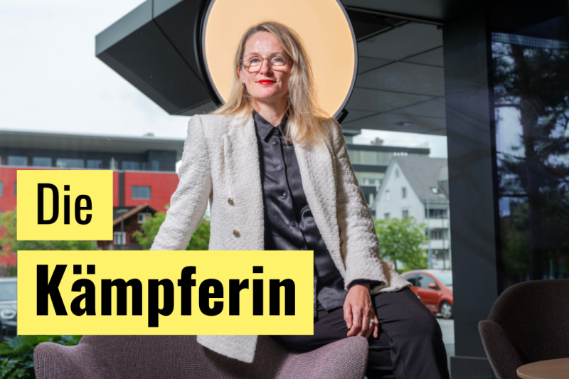 Grafische Anzeige mit der Überschrift „Die Kämpferin“ in schwarzer Schrift auf zwei knallgelben Rechtecken im linken Bildbereich. Im Hintergrund sitzt eine Frau mit Brille, rotem Lippenstift und weißem Tweed-Blazer auf der Armlehne eines mauvefarbenen Sessels. Hinter ihr leuchtet eine große kreisförmige Ringleuchte vor einer Glasfront mit Blick ins Freie.