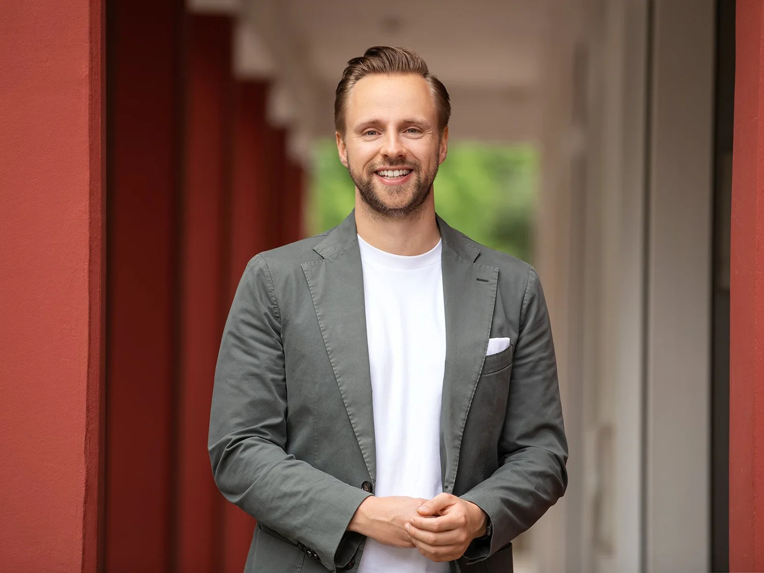 Ein freundlich lächelnder Mann mit kurzen braunen Haaren und Dreitagebart schaut direkt in die Kamera. Er trägt ein weißes T-Shirt unter einem grauen Sakko mit weißem Einstecktuch und hält die Hände locker vor dem Körper zusammengesteckt. Er steht in einem Säulengang mit terrakottafarbenen Wänden und verschwommenem Hintergrund.