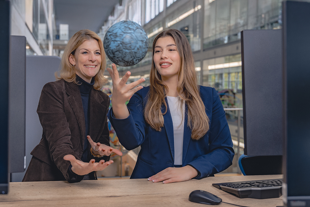 Zwei Frauen in Businesskleidung sitzen an einem Schreibtisch in einem modernen Büro mit grossen Fenstern. Eine Frau balanciert lächelnd einen Handball mit blauer Marmorierung auf der Fingerspitze, während die andere Frau daneben steht und den Ball mitgestikuliert. Auf dem Tisch befinden sich eine Tastatur und eine Computermaus, im Vordergrund sind Monitorränder sichtbar.
