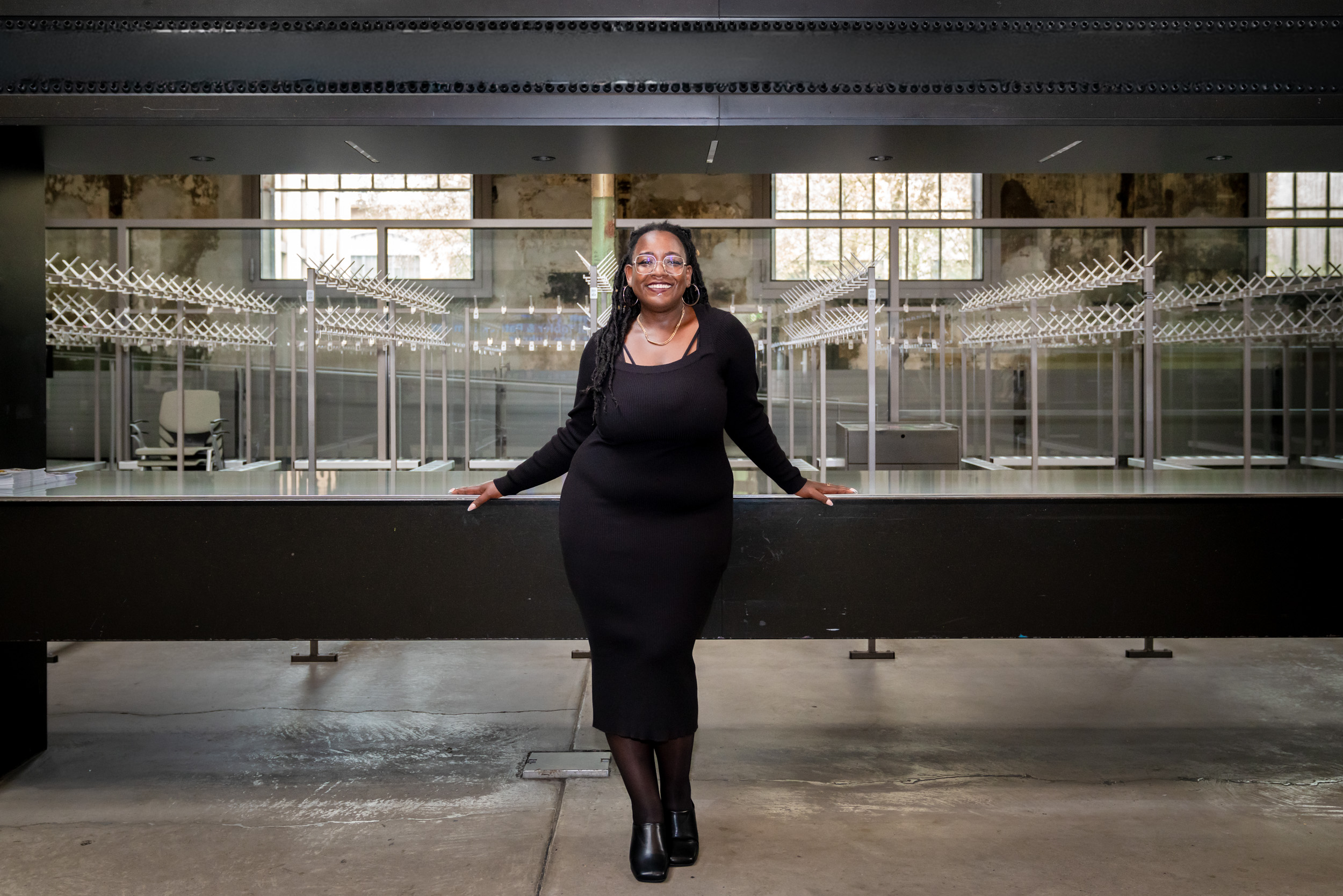 Yuvviki Dioh steht vor der leeren Garderobe im Schiffbau und lehnt an leicht an den Tresen an, ihre Arme nach aussen ausgebreitet,. Sie trägt ein schwarzes Kleid, Strümpfe und schwarze Schuhe, eine Brille und lächelt breit.