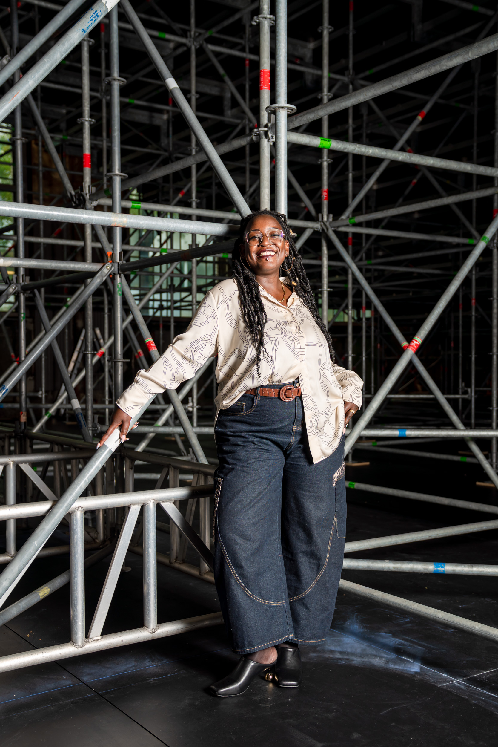 Yuvviki Dioh steht hinter der Schiffbau-Bühne, vor einem Gewirr aus Gerüsten, an die sie sich leicht anlehnt. Sie trägt blaue Jeans, einen braunen Gurt und eine hellbeige Bluse mit Knotenmuster. Sie lächelt breit in die Kamera.