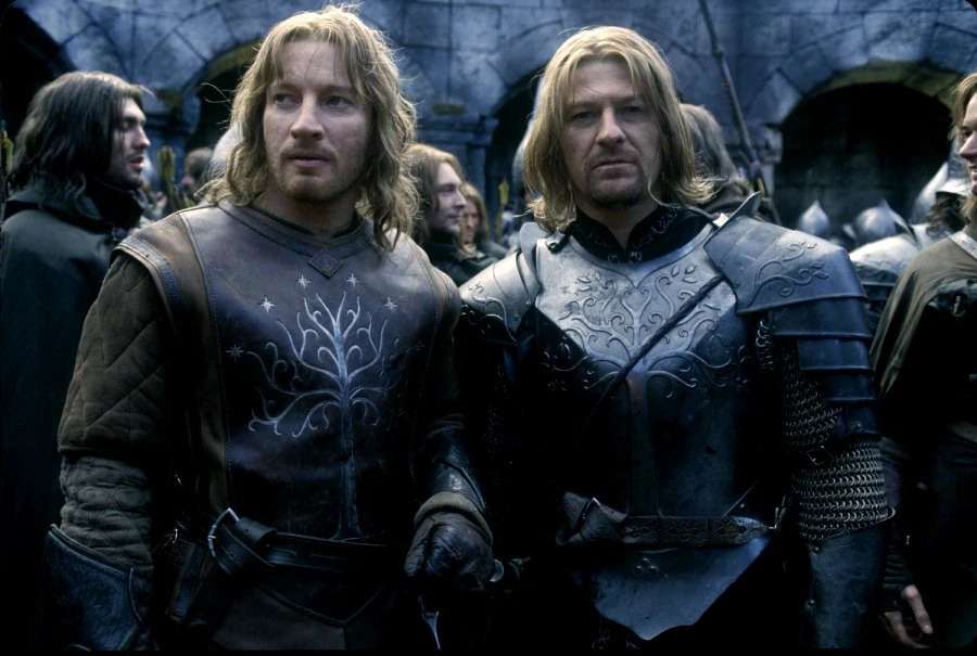 Ein Standbild aus einer Rückblende in der Extended Edition von „Der Herr der Ringe: Die zwei Türme“. Die Szene spielt in den grauen, steinernen Ruinen der umkämpften Stadt Osgiliath. Die Brüder Boromir und Faramir, beide in den Rüstungen Gondors, stehen dicht beieinander. Boromir lächelt stolz, hat liebevoll den Arm um seinen jüngeren Bruder gelegt und hält triumphierend die Standarte Gondors in die Höhe. Faramir blickt glücklich und voller Bewunderung zu seinem älteren Bruder auf. Im Hintergrund sieht man jubelnde Soldaten von Gondor, die ihren Sieg feiern. Die Stimmung zwischen den Brüdern ist warm, innig und voller gegenseitigem Respekt.