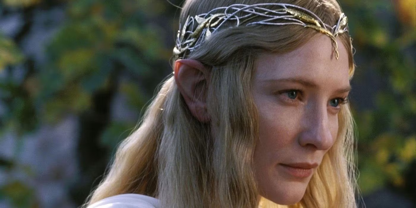 Eine detaillierte Nahaufnahme der Elbenherrin Galadriel, gespielt von Cate Blanchett. Sie ist im Halbprofil zu sehen und blickt mit einem sanften, weisen und fast etwas wehmütigen Ausdruck aus strahlend blauen Augen nach rechts aus dem Bild heraus. Sie hat langes, goldenes, leicht welliges Haar, durch das ihre spitzen Elbenohren deutlich sichtbar sind. Auf dem Kopf trägt sie ein sehr feines, filigranes Diadem, das aus ineinander verschlungenen silbernen Ranken und kleinen goldenen Blättern besteht. An ihrer linken Schulter ist der Ansatz eines fließenden weißen Gewandes zu erkennen. Der unscharfe Hintergrund zeigt weiches, warmes Sonnenlicht, das durch grüne und gelbliche Blätter fällt.
