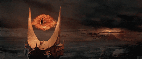 Ein animiertes GIF aus „Der Herr der Ringe“. Das riesige, flammende Auge Saurons mit seiner schmalen, katzenartigen Pupille schwebt zwischen den zwei großen, hornartigen Spitzen des dunklen Turms Barad-dûr. Das Auge knistert vor feuriger Energie und blickt langsam suchend hin und her. Im Hintergrund erstreckt sich die düstere, wolkenverhangene Landschaft von Mordor, in der rechts in der Ferne der Schicksalsberg rot glüht.