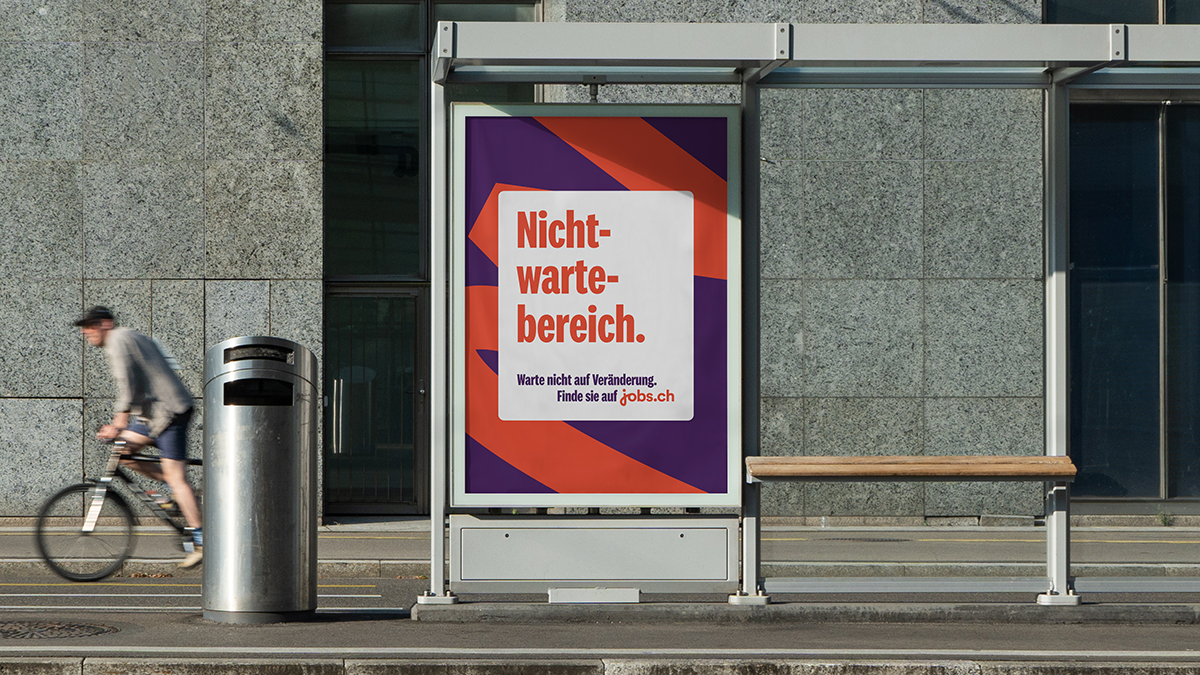 Ein Werbeplakat von jobs.ch in einem gläsernen Wartebereich an einer Straße. Das Plakat zeigt fettgedruckten, roten Text auf weißem Grund mit der Aufschrift „Nicht-warte-bereich.“. Darunter steht in kleinerer Schrift: „Warte nicht auf Veränderung. Finde sie auf jobs.ch“. Der Hintergrund des Plakats ist in dunklem Violett mit diagonalen orangenen Streifen gestaltet. Links im Bild fährt eine Person auf einem Fahrrad vorbei, im Hintergrund ist eine graue Steinfassade zu sehen.