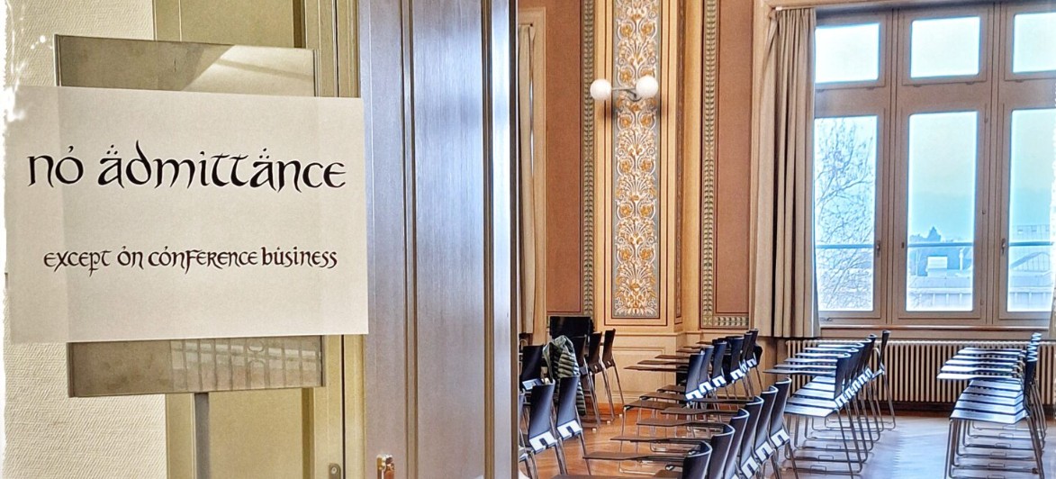 Das Bild zeigt den Blick durch eine geöffnete Tür. Im Vordergrund auf der linken Seite ist ein weisses Papierschild an einer Scheibe angebracht. Darauf steht in einer an das "Herr der Ringe"-Universum erinnernden Schriftart: "no admittance" und darunter etwas kleiner "except on conference business ". Viele Buchstaben sind dekorativ mit Akzenten oder Umlauten versehen. Der Spruch ist eine humorvolle Abwandlung des Schildes an Bilbo Beutlins Tür ("No admittance except on party business"). Auf der rechten Bildhälfte blickt man durch den hölzernen Türrahmen in einen grossen Konferenzraum in einem älteren, herrschaftlichen Gebäude. Die Wände sind mit klassischen, floralen Ornamenten verziert und eine runde Wandlampe ist zu sehen. Im Raum stehen ordentlich aufgereihte, leere moderne Konferenzstühle in Schwarz, die mit kleinen seitlichen Schreibflächen ausgestattet sind. Hohe Fenster lassen helles Tageslicht in den Raum fallen.