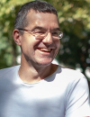 Ein freundlich lächelnder Mann mit Brille und kurzen, leicht ergrauten Haaren blickt leicht zur Seite. Er trägt ein weißes T-Shirt vor einem unscharfen, von Sonnenlicht durchfluteten grünen Hintergrund.