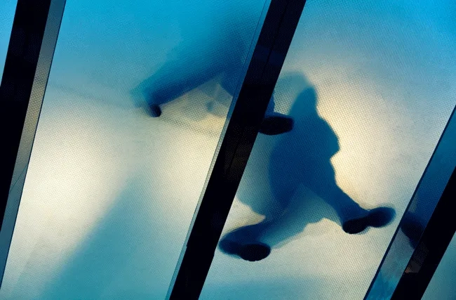 Zwei gehende Personen sind durch eine gläserne Decke von unten sichtbar, symbolisch für die Schwierigkeit für Frauen, Karriere zu machen und aufzusteigen.