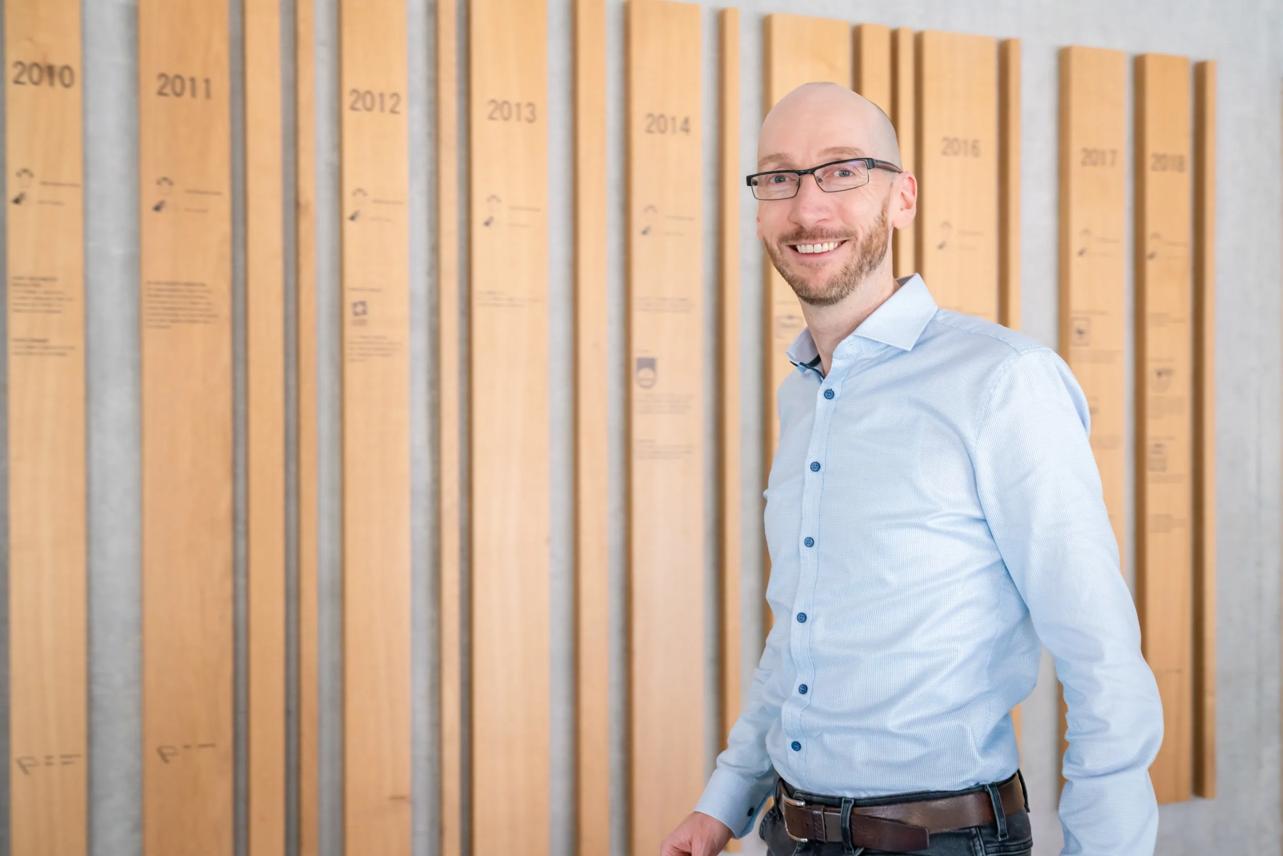 Das Bild zeigt Stefan Andexer, ein Mann mit Glatze, Hemd und Brille, sympathisch lachend vor der Lidl-History-Wand.