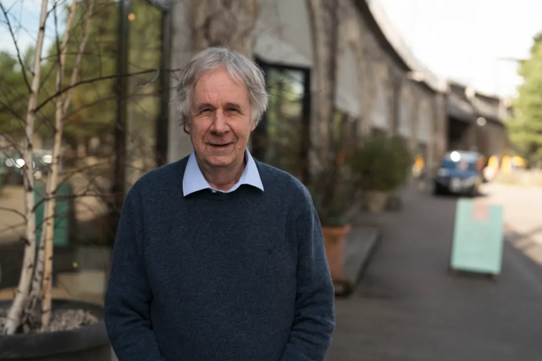 Das Bild zeigt François Höpflinger, ein älterer Mann mit dunkelblauem Pullover, darunter ein hellblaues Hemd, mit kurzen bis mittellangen Haaren. Er steht auf einem Trottoir, hinter ihm Strasse und Häuser.