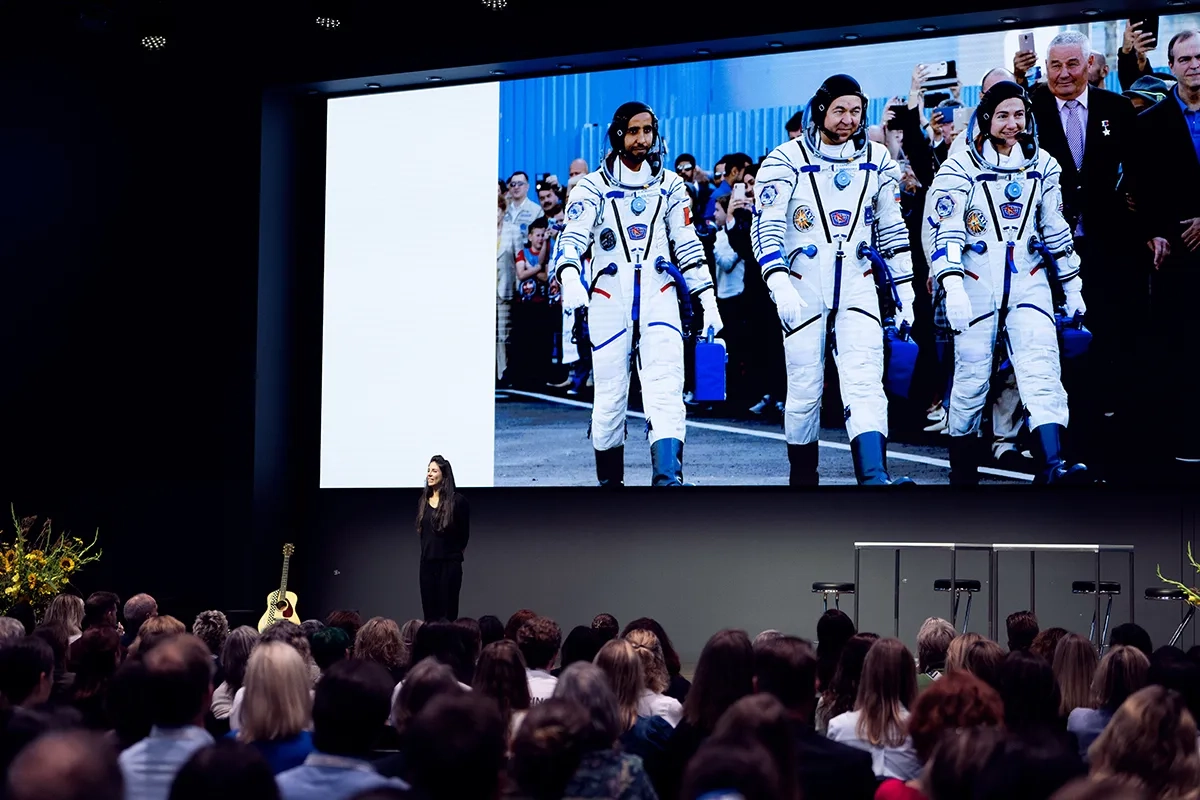Bühnenaufnahme eines Vortrags: Eine Referentin steht vor einem grossen Publikum; hinter ihr zeigt eine riesige Leinwand ein Foto von vier Astronauten und Astronautinnen in weissen Raumanzügen, die nebeneinander gehen. Links lehnt eine Gitarre, rechts stehen drei leere Hocker.