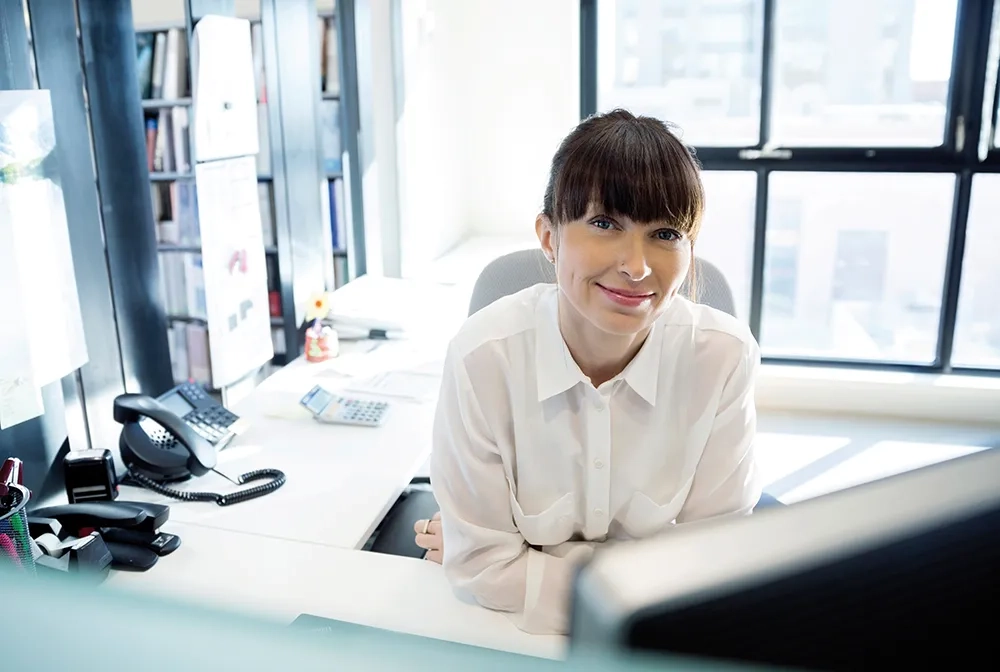 Person im weissen Hemd lächelt am Schreibtisch in einem hellen Büro; Telefon, Taschenrechner und Unterlagen liegen bereit.