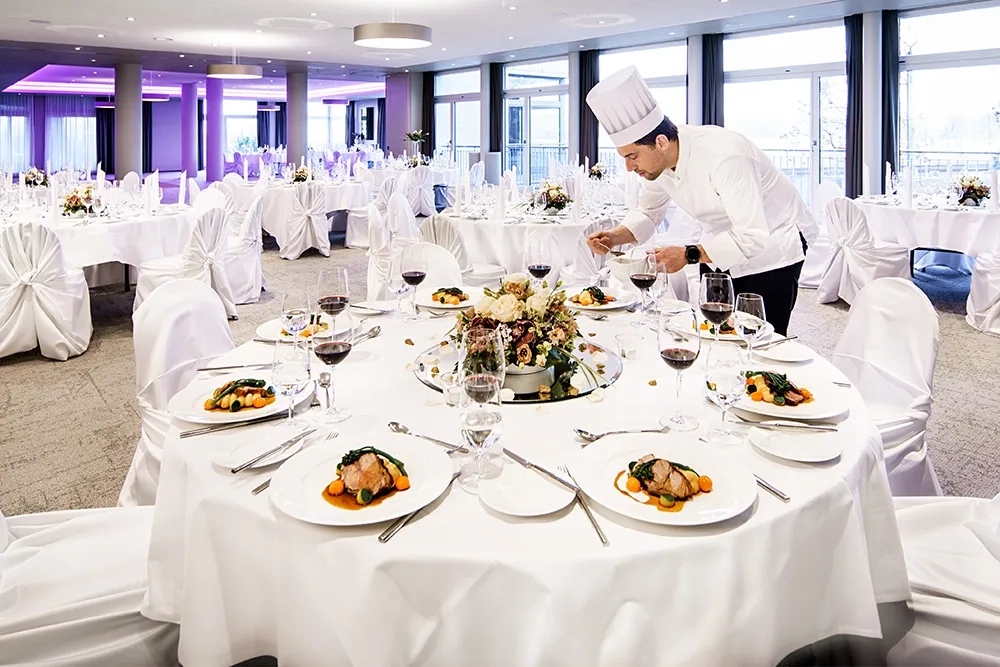 Eleganter Bankettsaal mit runden, weiss gedeckten Tischen und Stühlen mit weissen Hussen, festlich dekoriert mit Blumengestecken und Kerzen. Ein Koch in weisser Uniform richtet sorgfältig Teller auf einem gedeckten Tisch an. Die Atmosphäre ist hell und modern, mit lila Akzentbeleuchtung im Hintergrund und grossem Fensterfront mit Blick nach draussen.
