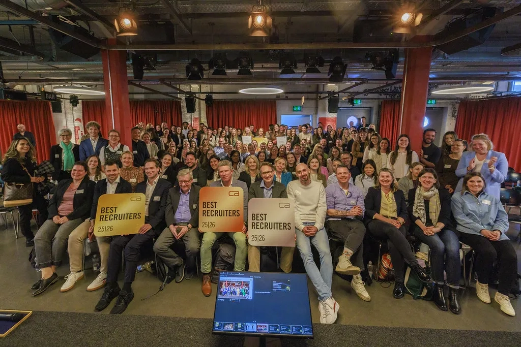 Eine große Gruppe von Menschen posiert für ein Gruppenfoto in einem Veranstaltungsraum im Industrie-Stil mit roten Vorhängen im Hintergrund. In der vorderen Reihe halten drei Personen große, quadratische Schilder mit der Aufschrift 'BEST RECRUITERS 25|26'. Im Vordergrund ist ein Monitor zu sehen, der die Aufnahmeumgebung eines Fotografen anzeigt.