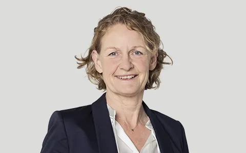 Ein Porträtfoto einer lächelnden Frau mit kurzen, lockigen, hellbraunen Haaren. Sie trägt einen dunkelblauen Blazer über einer weißen, leicht geöffneten Bluse. Sie trägt eine feine Halskette mit einem kleinen blauen Anhänger. Der Hintergrund ist durchgehend hellgrau.